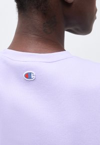 Κοντινό πλάνο στον λαιμό και το πάνω μέρος της πλάτης ενός ατόμου που φοράει ανοιχτό μωβ ριμπ μπλούζα με ένα σήμα Champion στο γιακά.