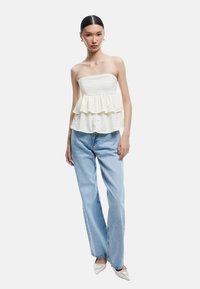Top crema sin tirantes con volantes en capas, combinado con jeans rectos de color azul claro. Modelo lleva tacones blancos puntiagudos. Diseño simple y texturizado.