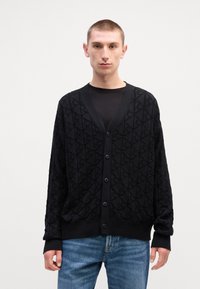 Mustan neulotun neuletakin, jossa on tekstuuri, V-pääntie, pitkät hihat ja napitus, yhdistettynä vaaleansinisiin farkkuihin.