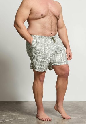 Mężczyzna bez koszulki, ubrany w kąpielówki w wzory, z rękami w kieszeniach, stojący boso na kamiennej podłodze przed gładką ścianą.