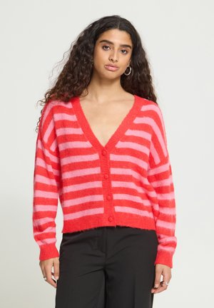 Donna con capelli lunghi e ricci che indossa un cardigan a righe rosso e rosa con bottoni e pantaloni neri su uno sfondo chiaro e uniforme.