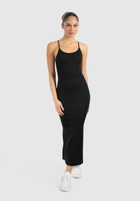 Vestido maxi negro, ajustado, con finas tirantes tipo espagueti, hecho de una tela elástica. La modelo lleva zapatillas blancas. Diseño simple y sin costuras.