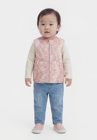 Taapero seisoo, pukeutuneena vaaleanpunaiseen kukkakuosiseen toppaliiviin, kermansävyiseen pitkähihainen paitaan, sinisiin farkkuihin ja vaaleanpunaisiin liukusukkiin.