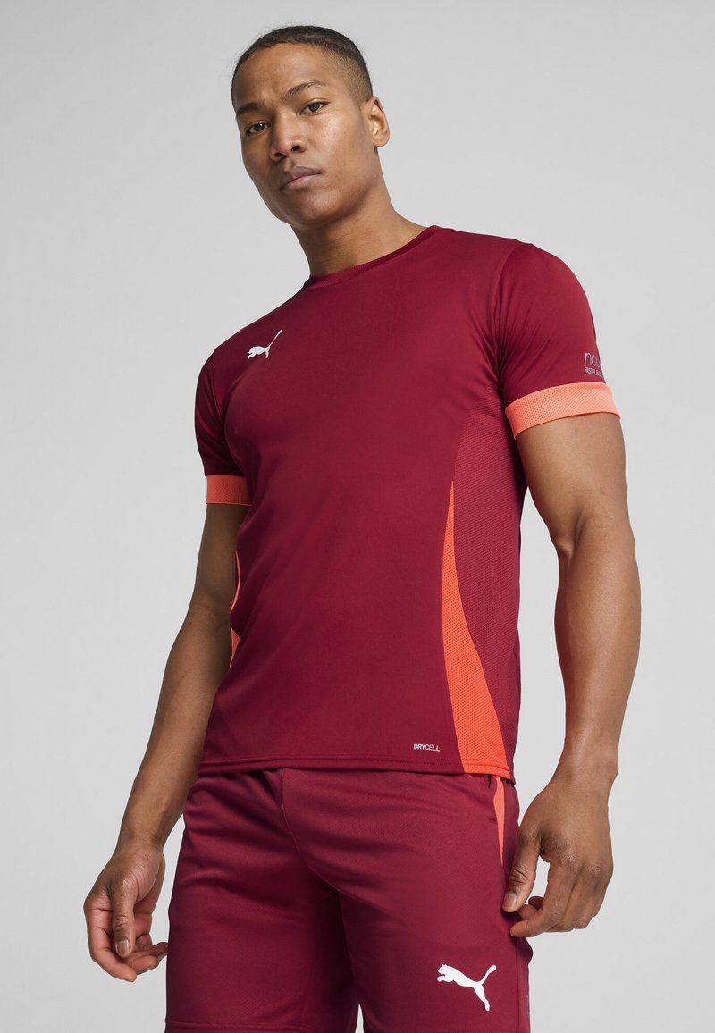 Bordeaux sportsjersey med orange accenter og kant. Fremstillet af et glat, fugttransporterende stof med et logo på brystet og siden.
