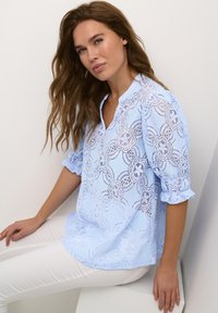 Blusa de encaje azul claro con patrones florales, escote redondo y mangas abullonadas. Combinada con pantalones skinny blancos.