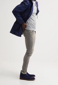 Giacca impermeabile navy sopra un maglione grigio, jeans grigio chiaro e stivaletti blu in camoscio con suole marroni. Taglio slim, stile casual.