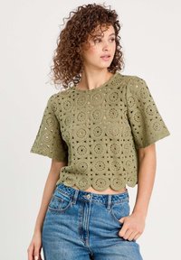 Haut crochet olive vert à manches courtes, avec un motif floral et un ourlet festonné, associé à un jean en denim bleu taille haute.