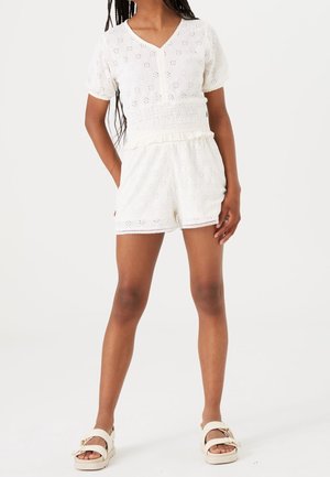 Pige iført hvid romper i øjlettet stof med korte ærmer og shorts samt hvide sandaler, stående med hænderne i lommerne.