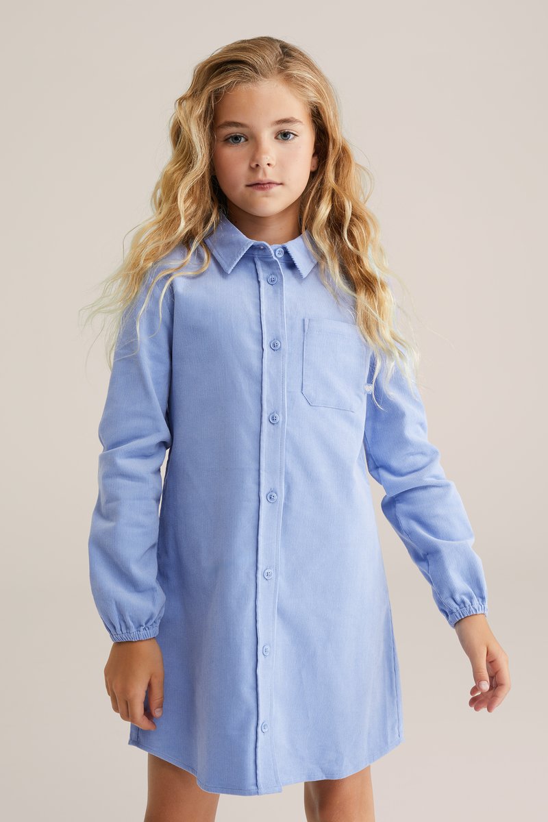 Rochie albastră cu mâneci lungi, prevăzută cu un buzunar pe piept. Confecționată dintr-un material moale cu o textură subtilă, cu manșete elastice și un tiv drept.