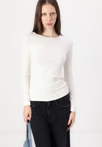 Langærmet ribbet hvid top med rund hals, parret med mørke jeans med nitter og holder en blå taske.