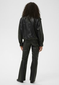 Personne aux cheveux bouclés portant une veste en cuir noire et un pantalon en cuir noir assorti, debout de dos sur un fond uni.