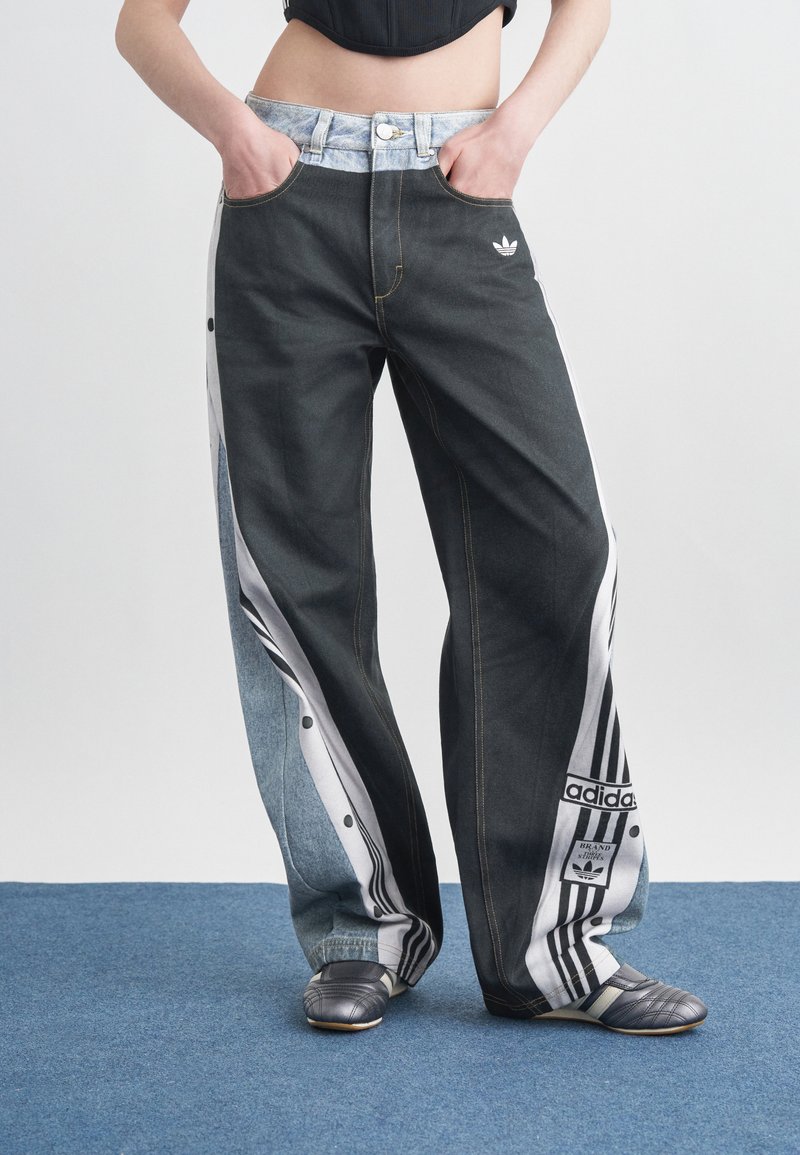 Άτομο που φοράει παντελόνι Adidas με φαρδιά μπατζάκια και συνδυασμό σκούρας και ανοιχτής τζιν ύφανσης, μαζί με μαύρες παντόφλες, στέκεται πάνω σε μπλε χαλί.