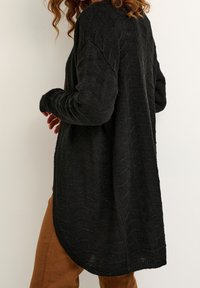 Kvinna som bär en texturerad svart stickad cardigan över bruna byxor, vänd något bort från kameran mot en neutral bakgrund.