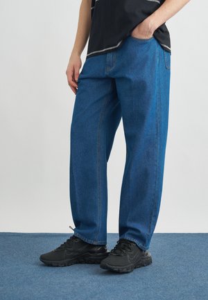 Személy laza kék farmert és fekete tornacipőt visel, egyik kezét a zsebében tartva áll kék szőnyegen.