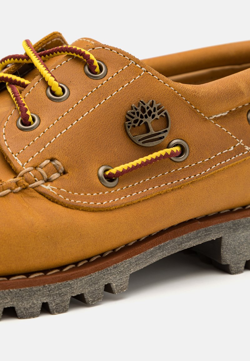 Bruine leren schoen met gele en kastanjebruine veters, een metalen logo-detail en een gestructureerde grijze rubberen zool met uitgesproken profiel.