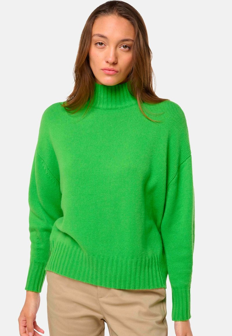 Femme portant un pull col roulé vert vif et un pantalon beige, se tenant devant un fond clair et uni.