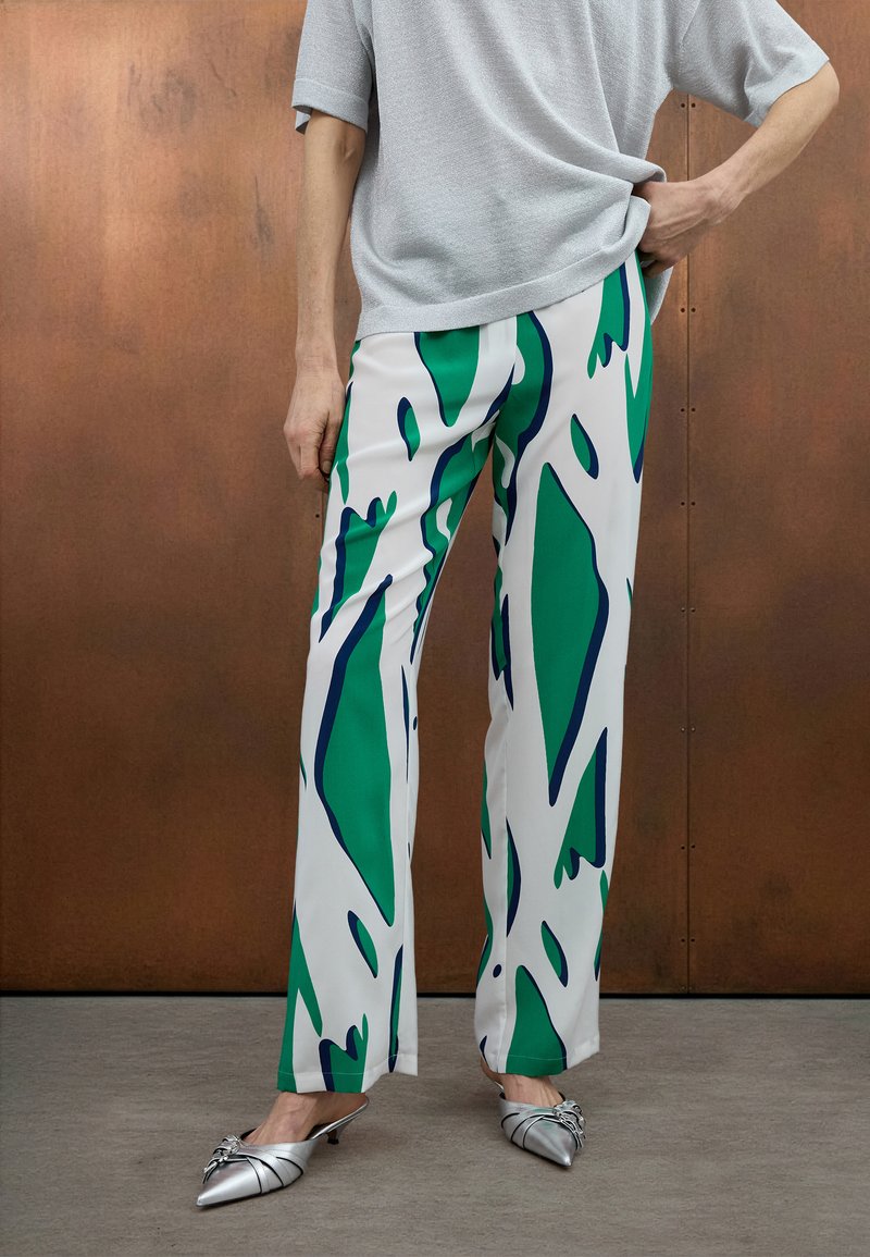 Pantalon blanc avec des motifs abstraits verts et bleu marine, associé à des mules à talons argentés. Coupe décontractée, texture lisse et longueur cheville.