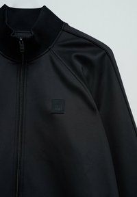 Chaqueta negra con cremallera hecha de textil suave; cuenta con un cuello alto y un parche cuadrado de logo en el pecho. Diseño simple y aerodinámico.