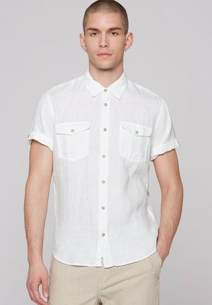 Mann mit kurzem Haar, der ein weißes kurzärmeliges Hemd mit zwei Brusttaschen und beigen Hosen trägt, steht mit einer Hand in der Tasche.