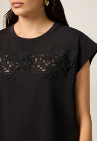 T-shirt nera in cotone con un design di ricamo floreale sul petto. Maniche corte con scollatura rotonda, dettaglio in tessuto testurizzato.