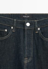Jeans in denim indaco scuro con cuciture dorate a contrasto, chiusura a bottoni e design a cinque tasche. Etichetta con la scritta "Massimo Dutti", realizzati in Marocco.