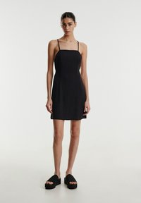 Schwarzes Minikleid mit dünnen Trägern, geradem Ausschnitt und glatter Textur. Kombiniert mit schwarzen Plateauschuhen zum Hineinschlüpfen.