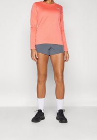 Koraalkleurig sportshirt met lange mouwen en een ronde halslijn, gecombineerd met grijze shorts. Zwarte sneakers en witte sokken maken de outfit af.