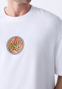 Hvid bomulds T-shirt med et rundt, farverigt "Duff" logo med orange, gule og sorte elementer, trykt i en fed, legende stil.