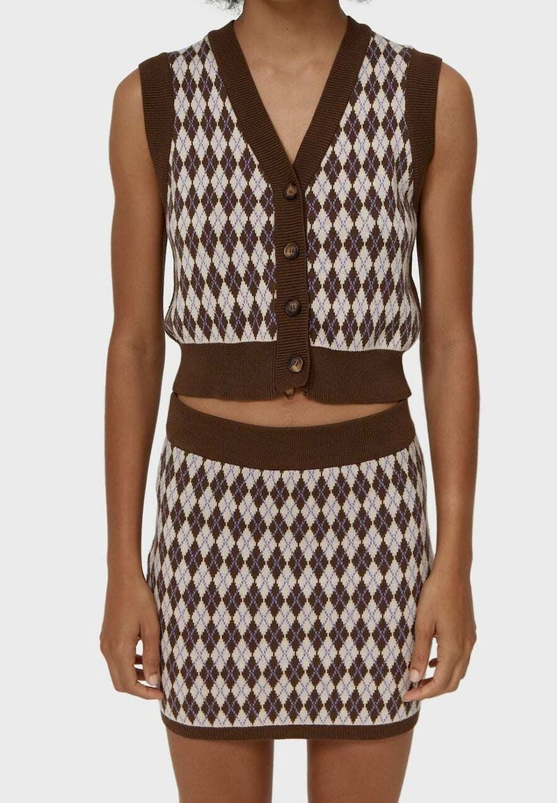 Gilet sans manches en tricot à motif losanges marron et beige avec des boutons, assorti à une mini-jupe taille haute en tricot à motif losanges, porté par une personne debout.