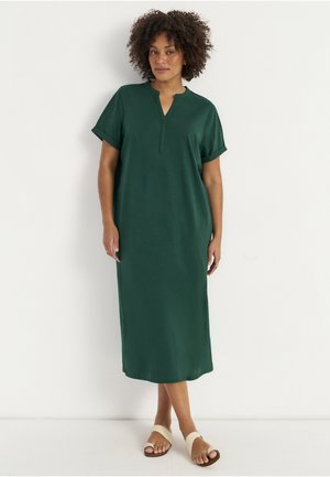 Kvinde med krøllet hår iført en mørkegrøn, kortærmet, knælængde kjole og beige slid-sandaler, der står mod en hvid baggrund.