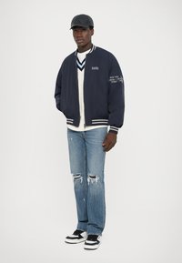 Jachetă de bombardier navy cu dungi albe, text grafic, pulover crem, blugi blue jeans deteriorați și adidași alb-negru. Căciulă neagră.