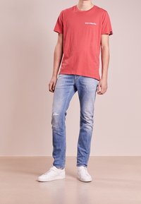 Person wearing a red "Have A Nice Day" t-shirt, light distressed jeans, and white sneakers, standing against a plain background.