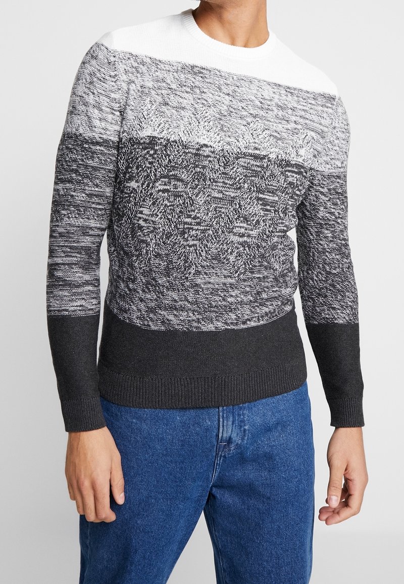 Homme portant un pull en maille à motifs noir, blanc et gris avec un jean bleu sur un fond blanc uni.