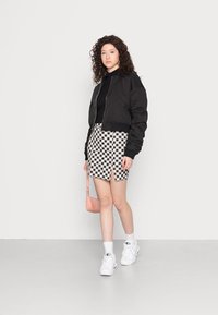 Corta bomber preta sobre uma blusa preta, combinada com uma saia mini xadrez com uma racha frontal. Ténis brancos volumosos e uma pequena bolsa rosa.