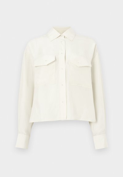 Chemise blanche courte en tissu lisse, avec col pointu, fermeture à boutons et deux poches poitrine avec rabats.