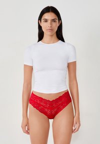 Top corto bianco aderente a maniche corte abbinato a mutandine rosse in pizzo a taglio ribassato, con motivi floreali e bordi a scallop lungo l'elastico.