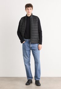 Colete acolchoado preto sobre um suéter preto, combinado com jeans azul claro e ténis pretos. O colete tem um fecho na frente e um emblema.