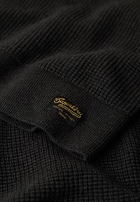 Suéter negro de punto hecho de tela acanalada, con un patrón texturizado. Incluye una etiqueta de marca amarilla prominente con el logo "Superdry".