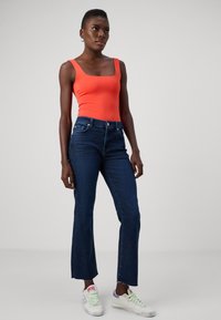 Rode body met vierkante hals, donkerblauwe flared jeans met rafelrand, witte sneakers met pastelaccenten, model staande tegen een lichte achtergrond.
