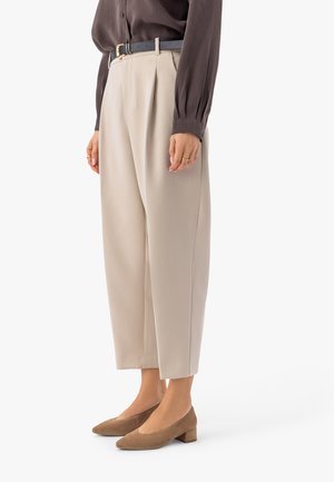 Kobieta ubrana w beżowe, krótkie spodnie z wysokim stanem, ciemny pasek, brązową bluzkę z długim rękawem oraz brązowe buty na niskim obcasie, stojąca.