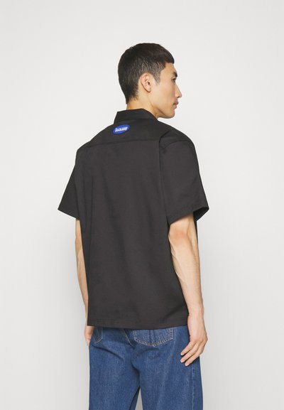 Melna īsām piedurknēm krekls no gludas auduma, ar norādītu apkakli, taisnu piegriezumu un zilu logo uzlīmi aizmugurē.