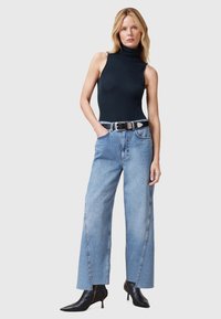 Top negro sin mangas con cuello alto, jeans de tiro alto de piernas anchas en azul claro, cinturón negro con detalles plateados y botas negras de tacón puntiagudas.
