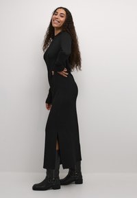 Black long-sleeve dress with a side slit, made from textured fabric. Paired with knee-high black boots featuring buckle accents.