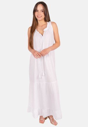 Frau mit langen braunen Haaren, die ein ärmelloses weißes Maxikleid mit Quasten und beigen Sandalen mit Absatz trägt, lächelt in die Kamera.