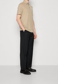 Man wearing beige short-sleeve polo shirt, black wide-leg trousers, and black loafers standing with hands in pockets.