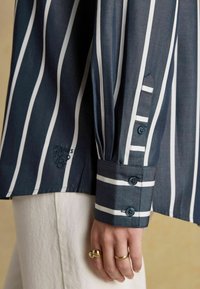 Tumesinine ja valge vertikaalsete triipudega pikkade varrukatega särk, nööpidega mansett, brodeeritud logo, valged püksid ning käsi kuldrõngastega.