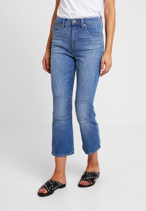 Vaqueros de denim azul claro con cintura alta, piernas ligeramente acampanadas y detalles de parches en las rodillas. Combinados con sandalias deslizantes negras con tachuelas.