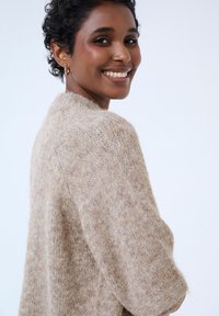 Beige knitted sweater with a relaxed fit, featuring a high neckline and textured, soft fabric. Long sleeves and minimal detailing.