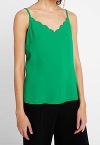 Camisola verde con scollatura a onda, spalline sottili e texture liscia. Abbinata a pantaloni neri.
