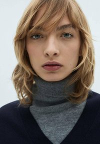 Blonde lase s softimi valovi, oblečena v temno mornarsko modro pulover s V-izrezom nad sivo polo majico. Svetla koža, nevtralni make-up in modre oči so vidni.